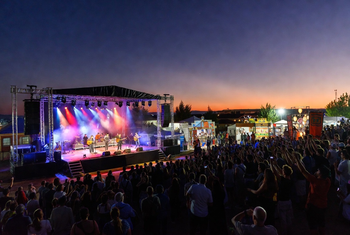 A festival stage lit at sunset with a crowd, raised hands, and food stalls along the edge