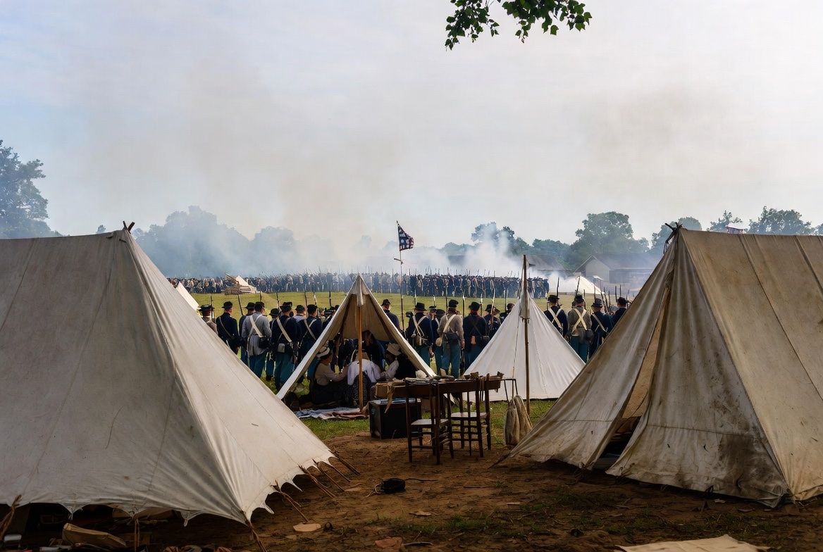 White canvas tents framing ranks of reenactors in formation under drifting musket smoke
