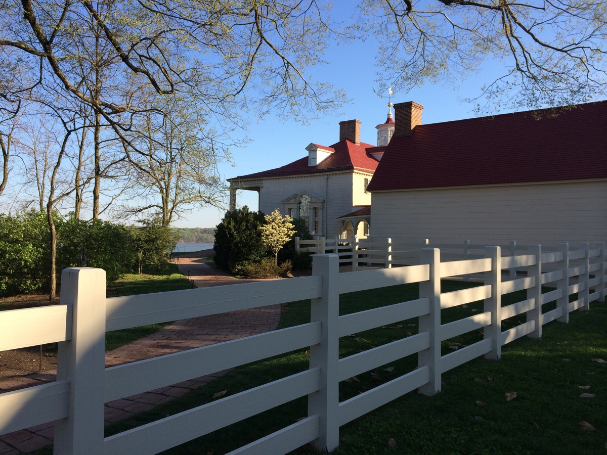 A white fence along a lane leading past a historic house toward the river