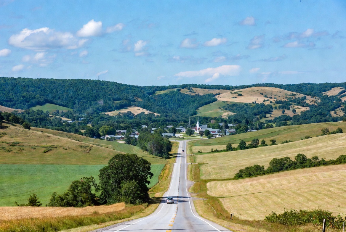 A two-lane highway rolling through farmland toward a small town with a church steeple